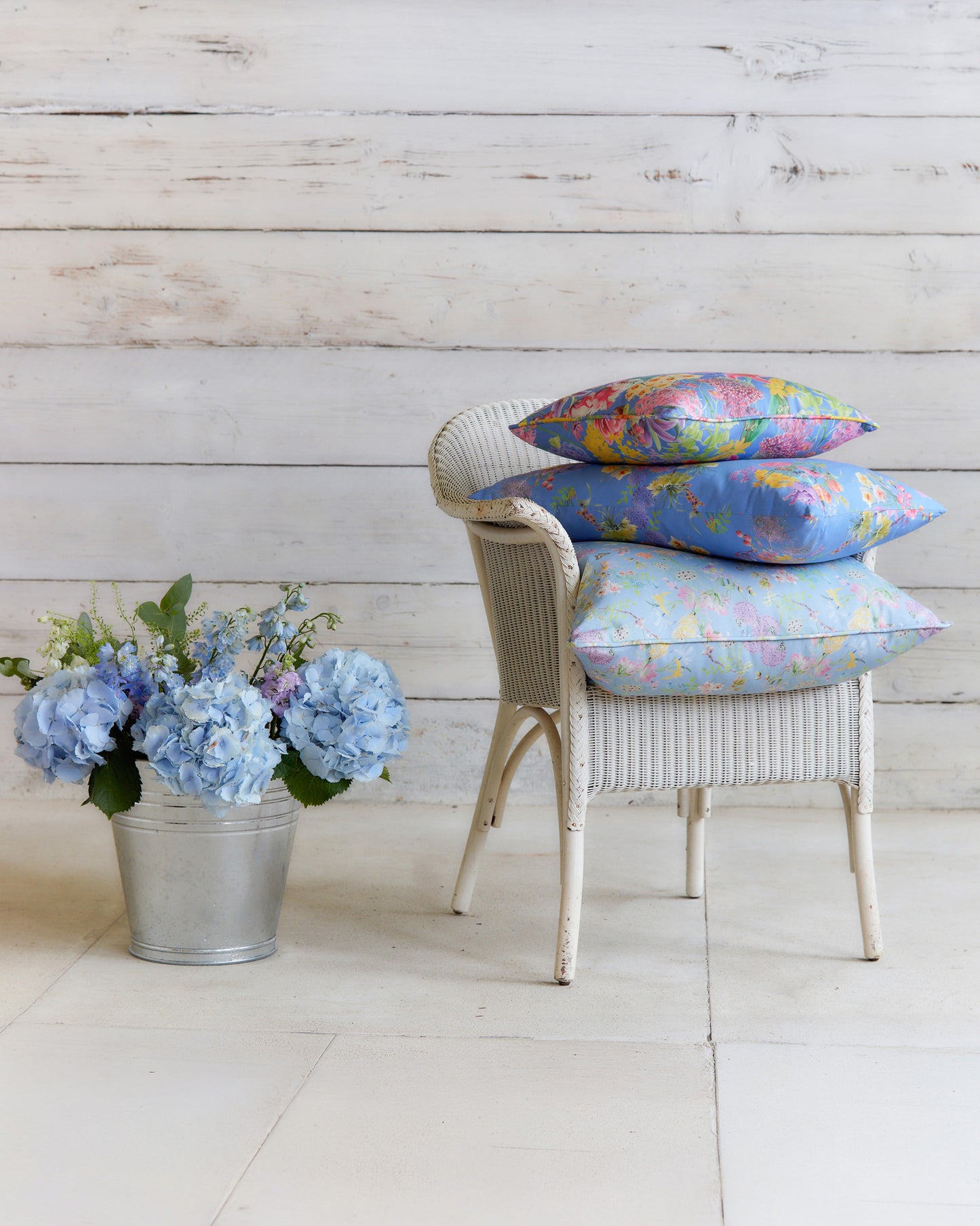Burst into Bloom Blue floral cushions by Bauldry Botanicals displayed on a white wicker chair, showcasing colourful botanical fabric for home interiors.
