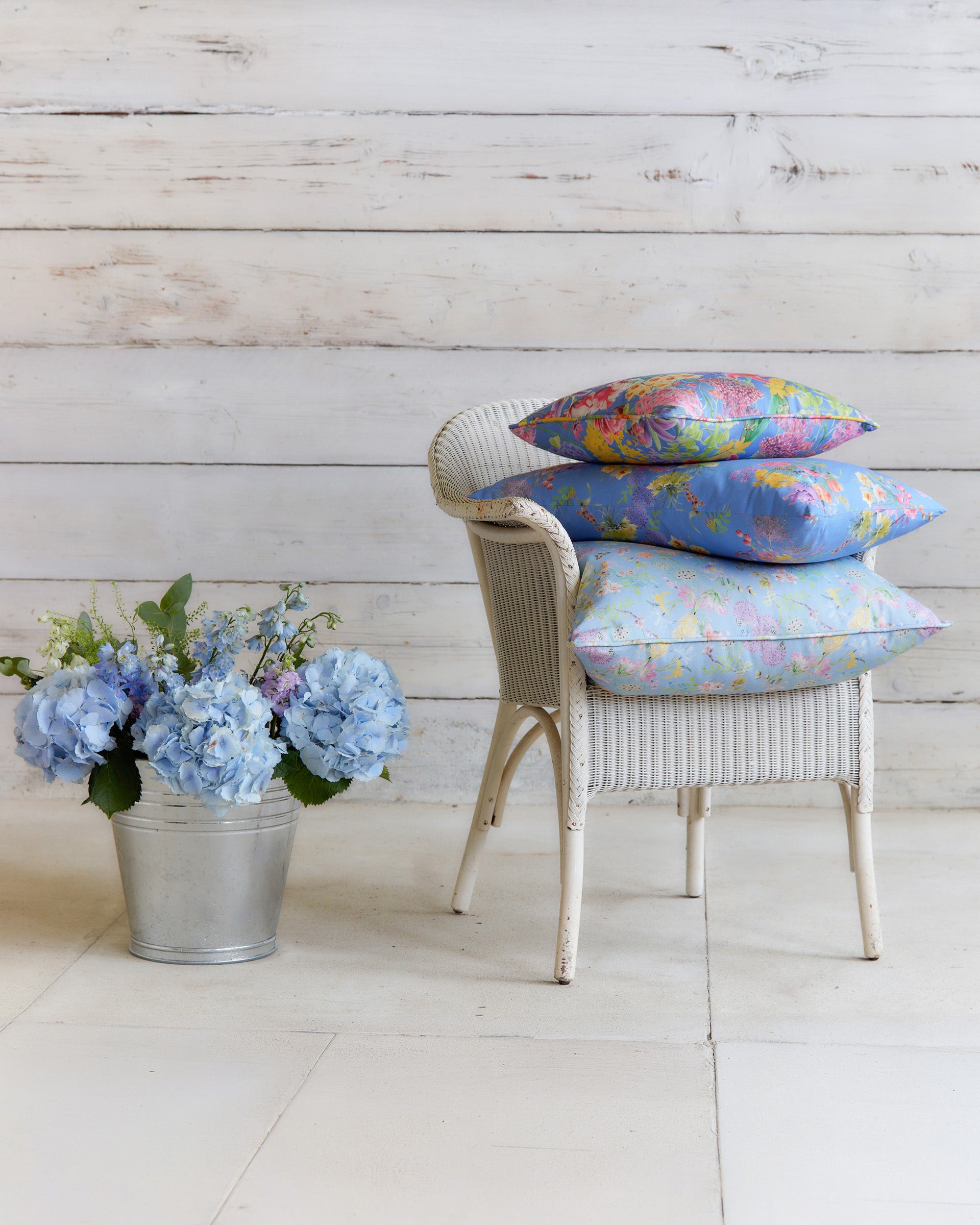Burst into Bloom Blue floral cushions by Bauldry Botanicals displayed on a white wicker chair, showcasing colourful botanical fabric for home interiors.