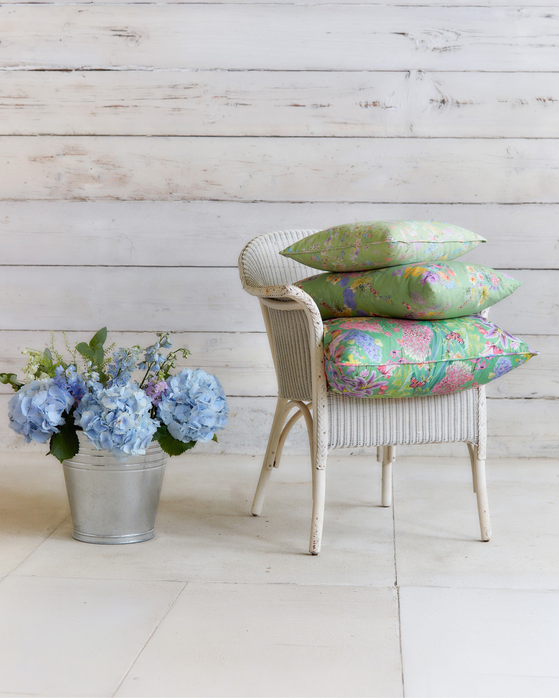 Burst into Bloom Green floral cushions by Bauldry Botanicals displayed on a white wicker chair, showcasing colourful botanical fabric for home interiors.