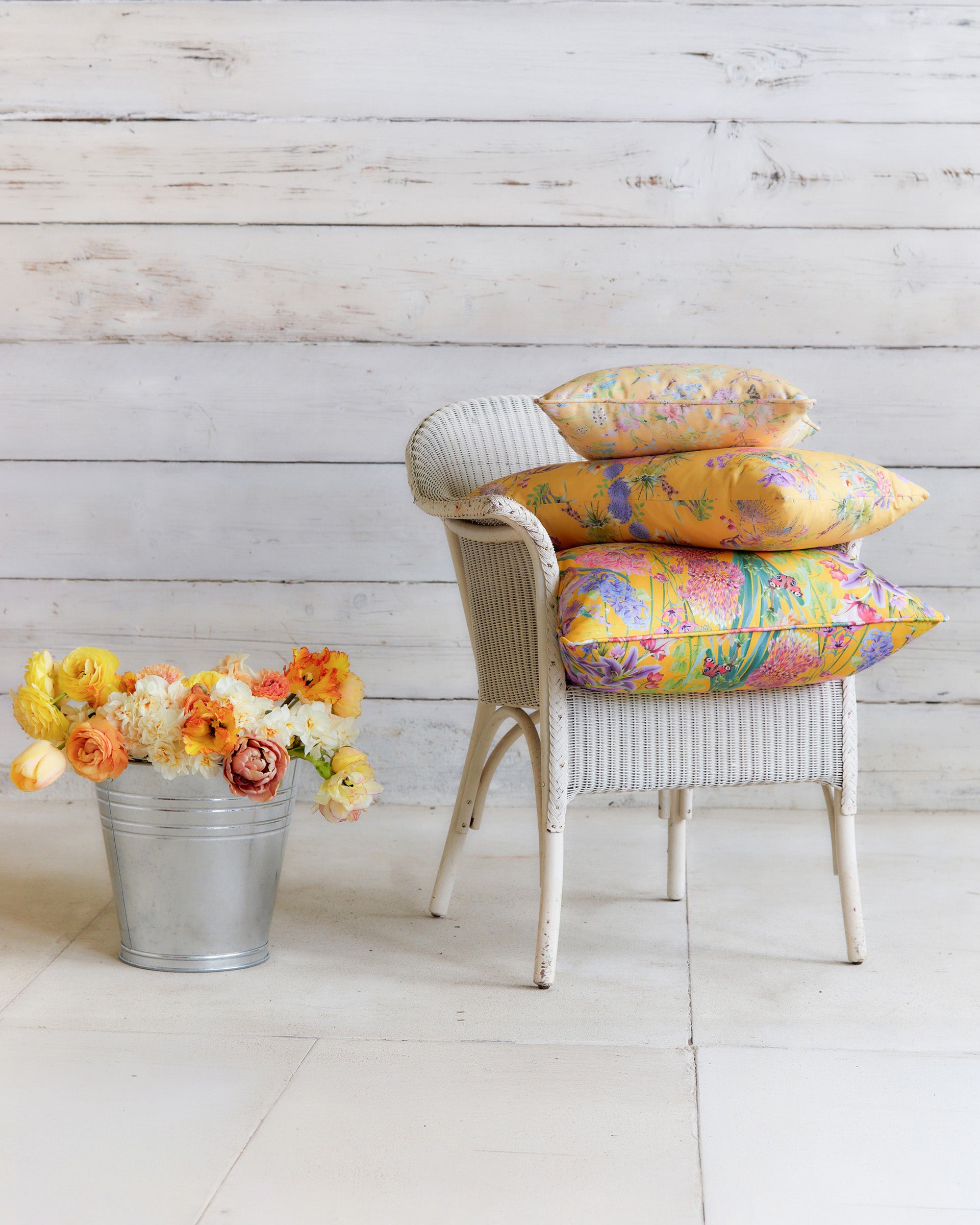 Burst into Bloom yellow floral cushions by Bauldry Botanicals displayed on a white wicker chair, showcasing colourful botanical fabric for home interiors.